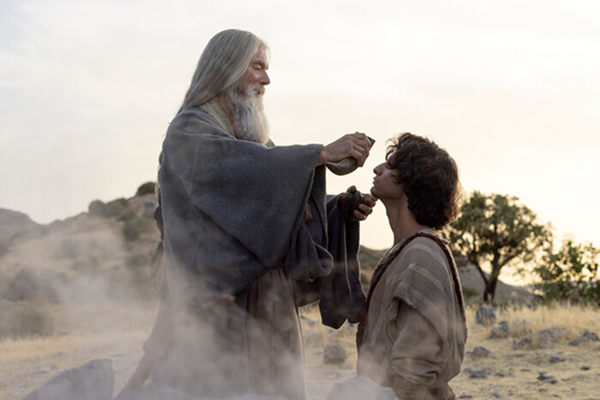 Outdoor scene with elder figure blessing younger character in dusty environment